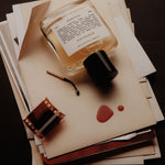 A clear glass perfume bottle labeled NOSTALGIA  with a black cap, arranged on top of stacked cream-colored papers and envelopes. A small film strip, a match, and two reddish-brown ink spots are also visible on the papers against a dark background.