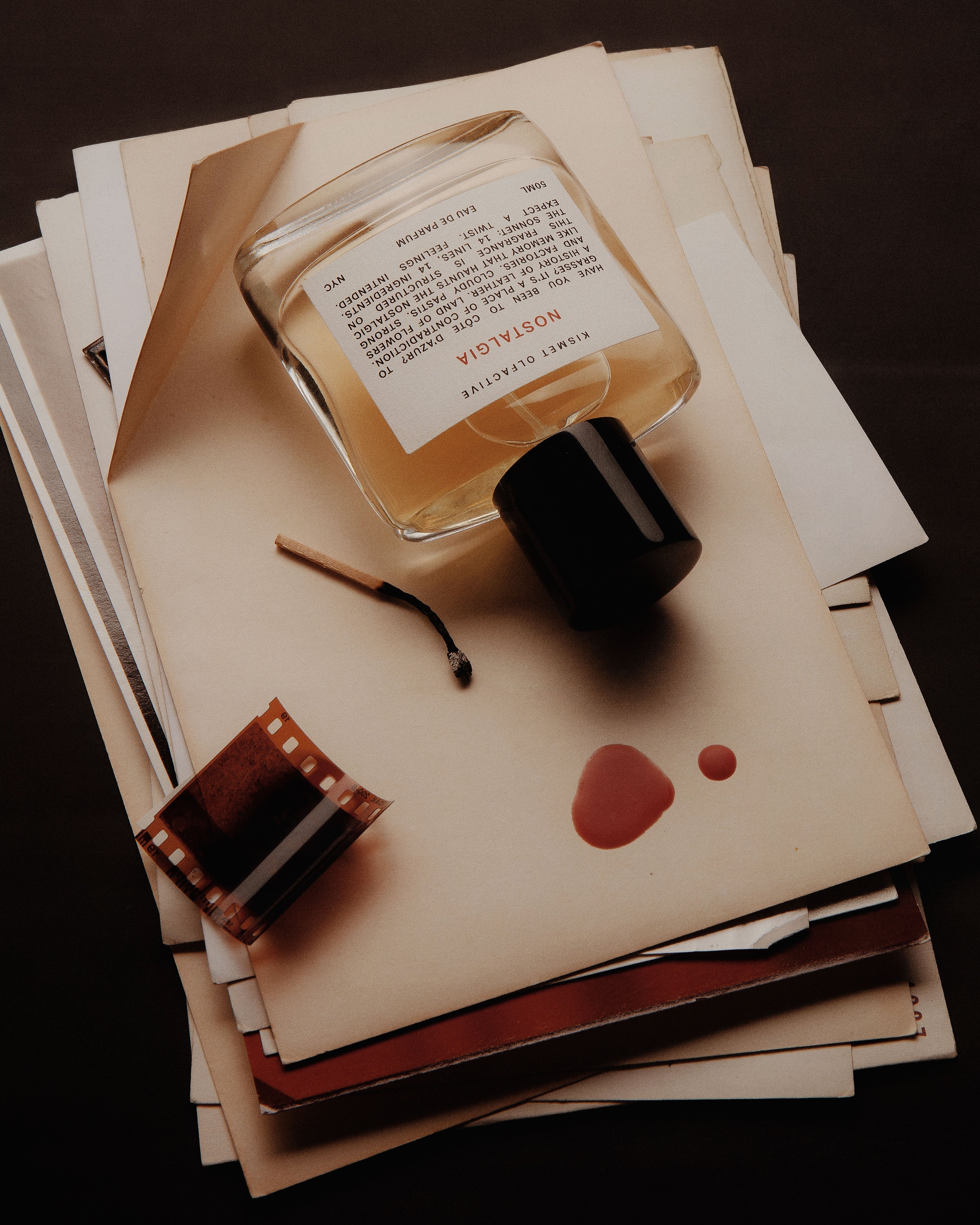 A clear glass perfume bottle labeled NOSTALGIA  with a black cap, arranged on top of stacked cream-colored papers and envelopes. A small film strip, a match, and two reddish-brown ink spots are also visible on the papers against a dark background.