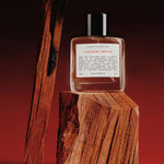 A clear glass perfume bottle labeled 'Kismet Olfactiev Tuberose Tantra' sits on a rough wooden block, with two tall burnt wood sticks leaning against a dark red background.
