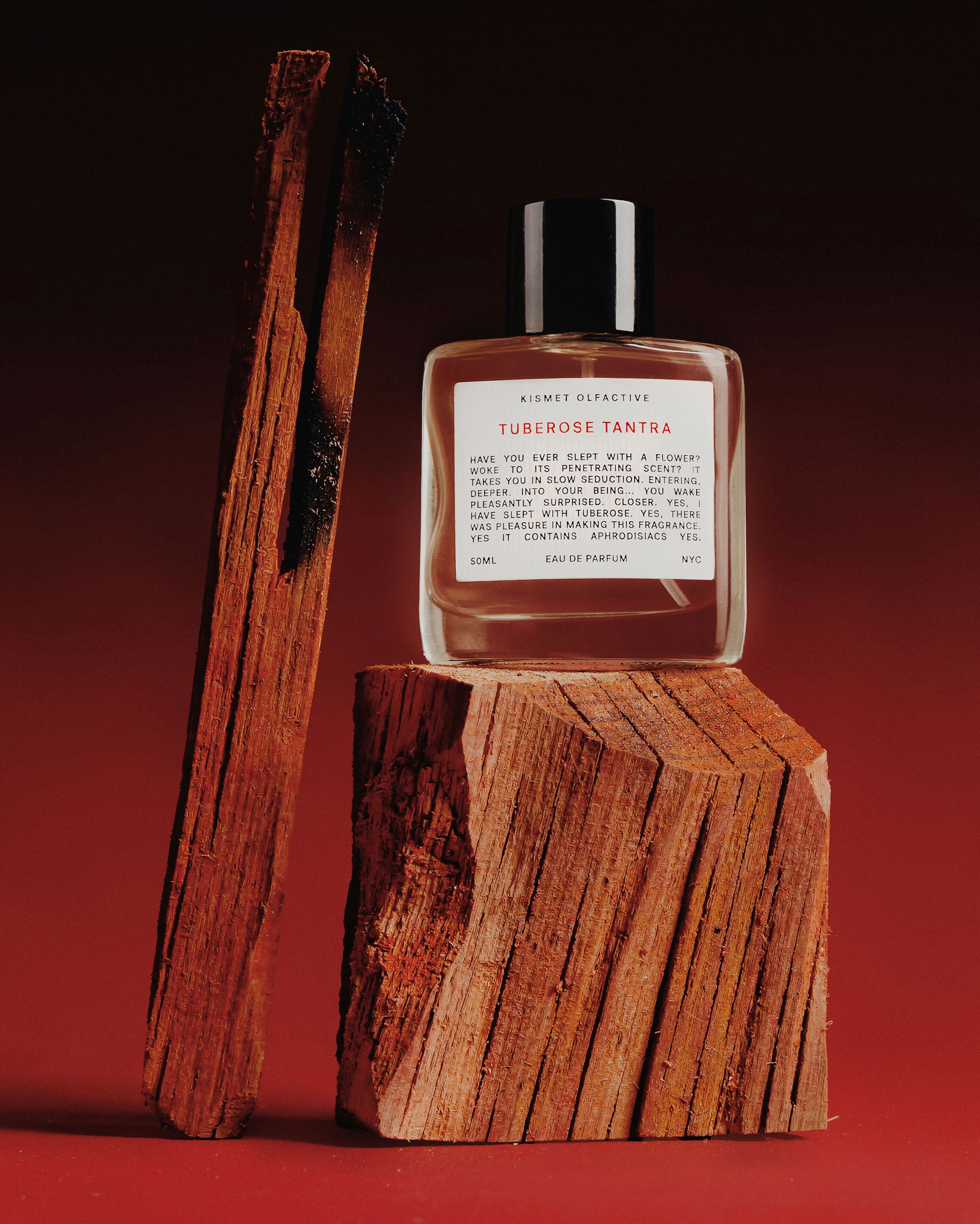 A clear glass perfume bottle labeled 'Kismet Olfactiev Tuberose Tantra' sits on a rough wooden block, with two tall burnt wood sticks leaning against a dark red background.