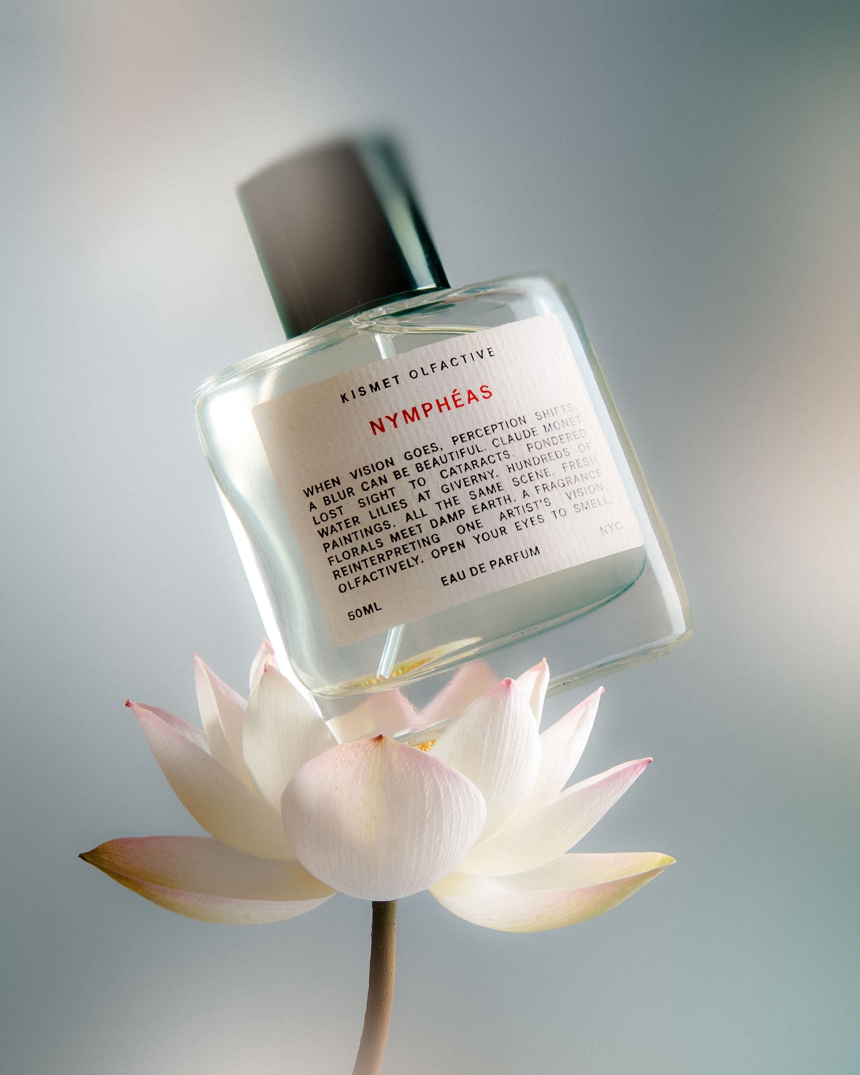 A clear glass perfume bottle labeled 'KIMSET OLFACTIVE Nymphéas' with a black cap, resting on a white lotus flower against a soft gray background.