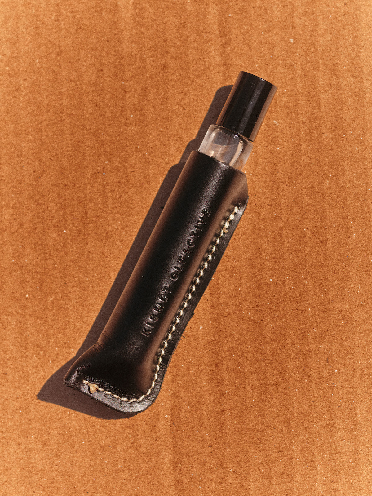 A small clear glass perfume bottle with a black cap, nestled inside a black leather sleeve with white stitching, placed diagonally on a textured brown cardboard background.