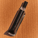 A small clear glass perfume bottle with a black cap, nestled inside a black leather sleeve with white stitching, placed diagonally on a textured brown cardboard background.