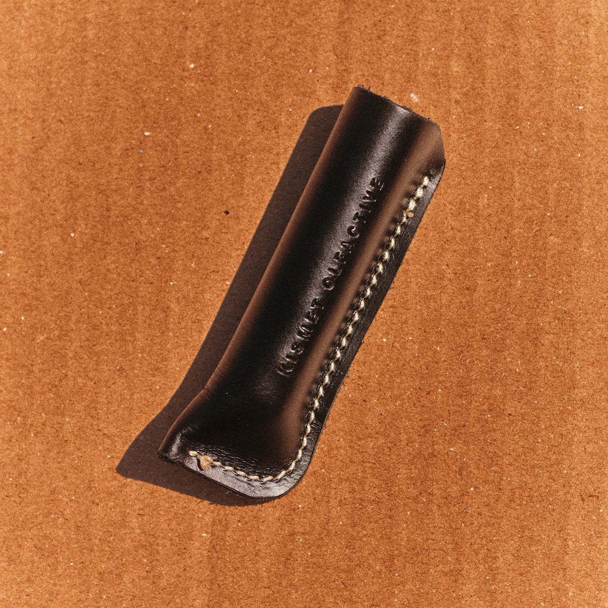 A dark leather perfume travel case with white contrast stitching and debossed branding, shown on a brown cardboard background.