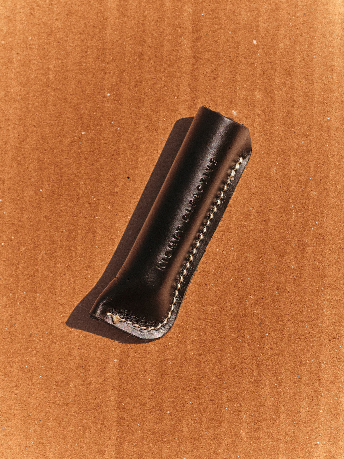 A dark leather perfume travel case with white contrast stitching and debossed branding, shown on a brown cardboard background.