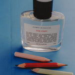 A clear glass perfume bottle with a black cap and a white label reading 'KISMET OLFACTIVE THE POET' on a blue background, with three colored pencils placed below it.