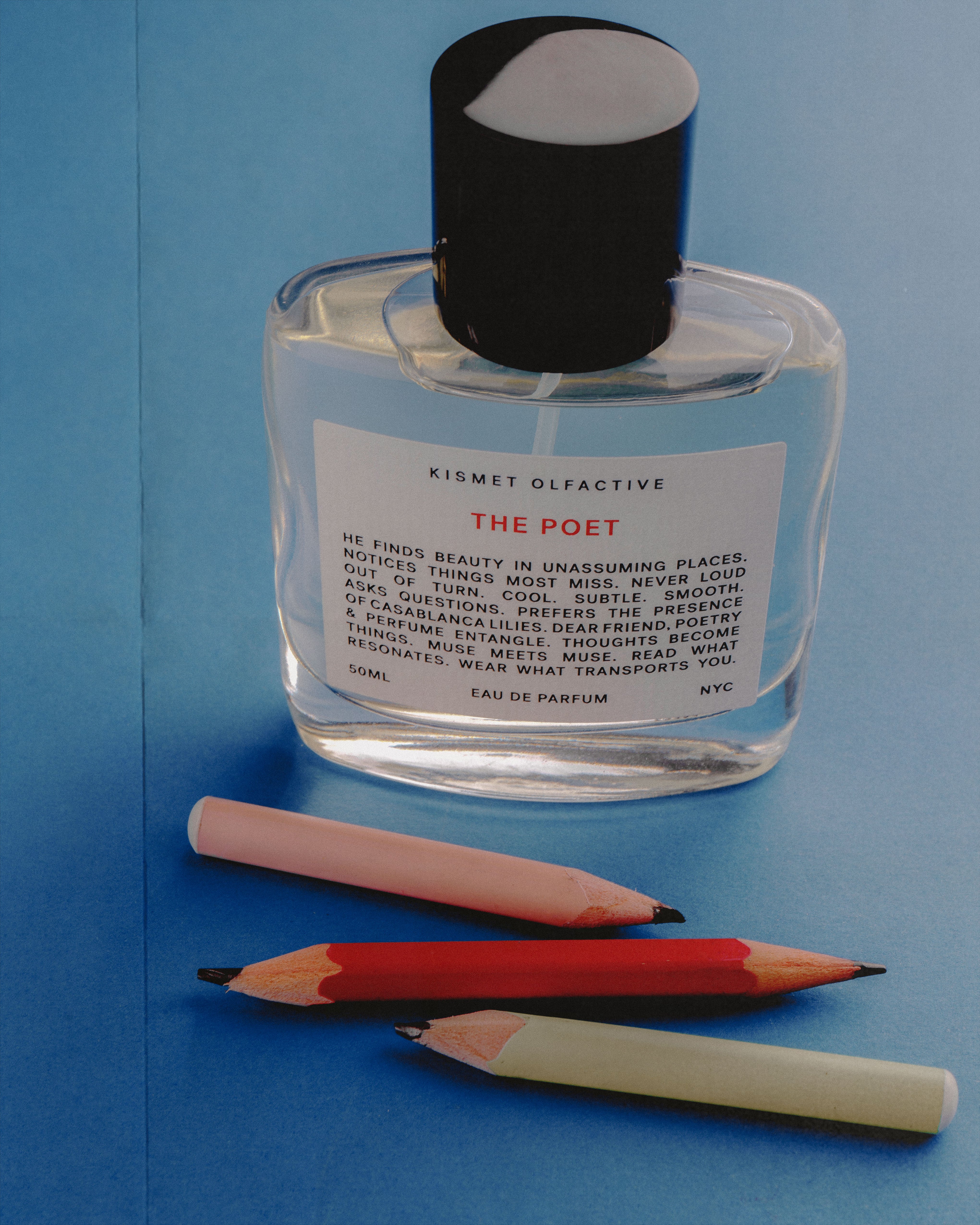 A clear glass perfume bottle with a black cap and a white label reading 'KISMET OLFACTIVE THE POET' on a blue background, with three colored pencils placed below it.