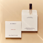 white kismet olfactive sample set box on a beige background and one perfume sample vial peeking out of box