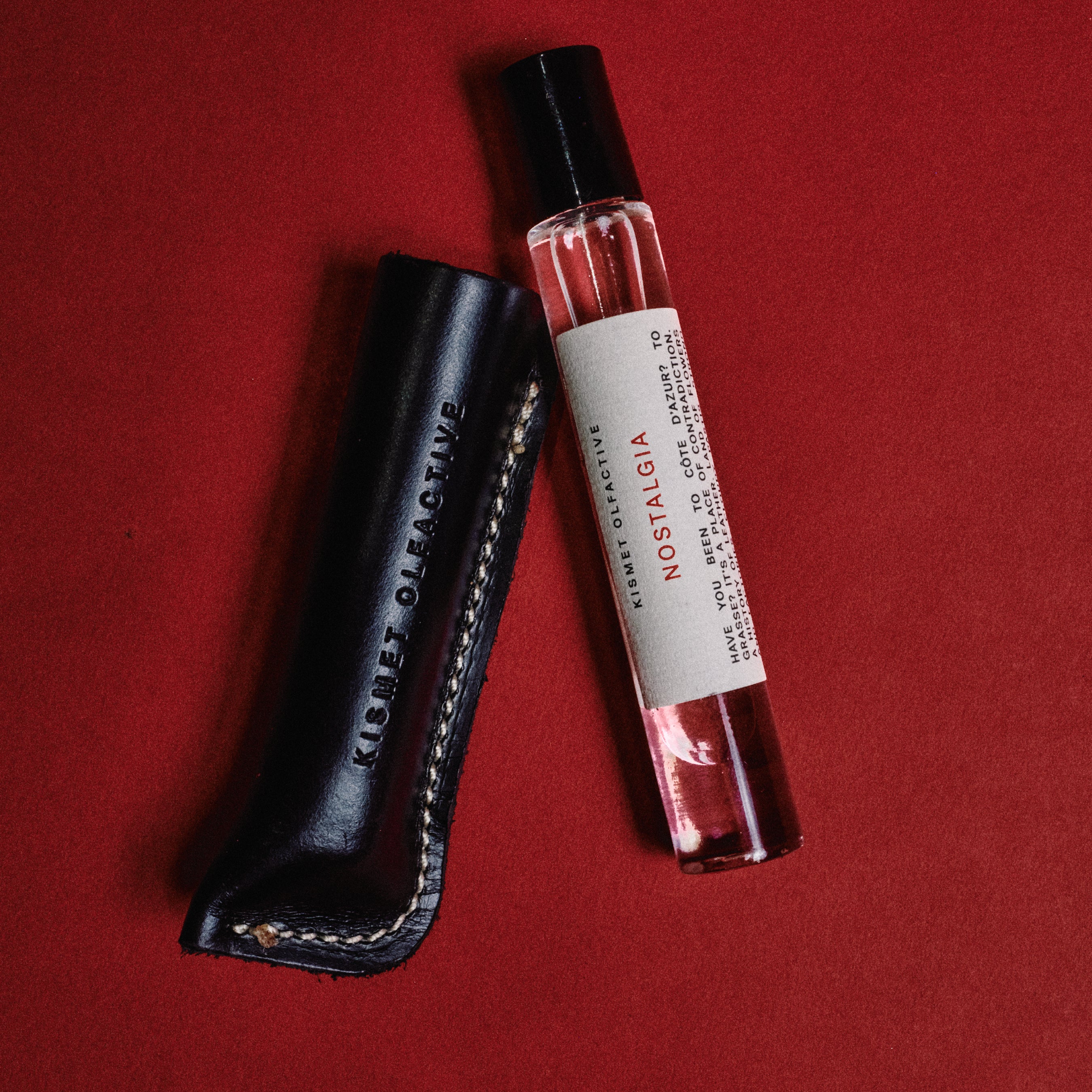 A small clear perfume or fragrance roller bottle with a white label reading NOSTALGIA, placed beside a black leather case embossed with  KISMET OLFACTIVE, on a solid red background.