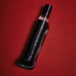 A black leather perfume bottle sleeve holding a clear glass perfume bottle with pink liquid, set diagonally on a textured red background. The sleeve features white contrast stitching and the brand name 'KISMET OLFACTIVE' debossed vertically.