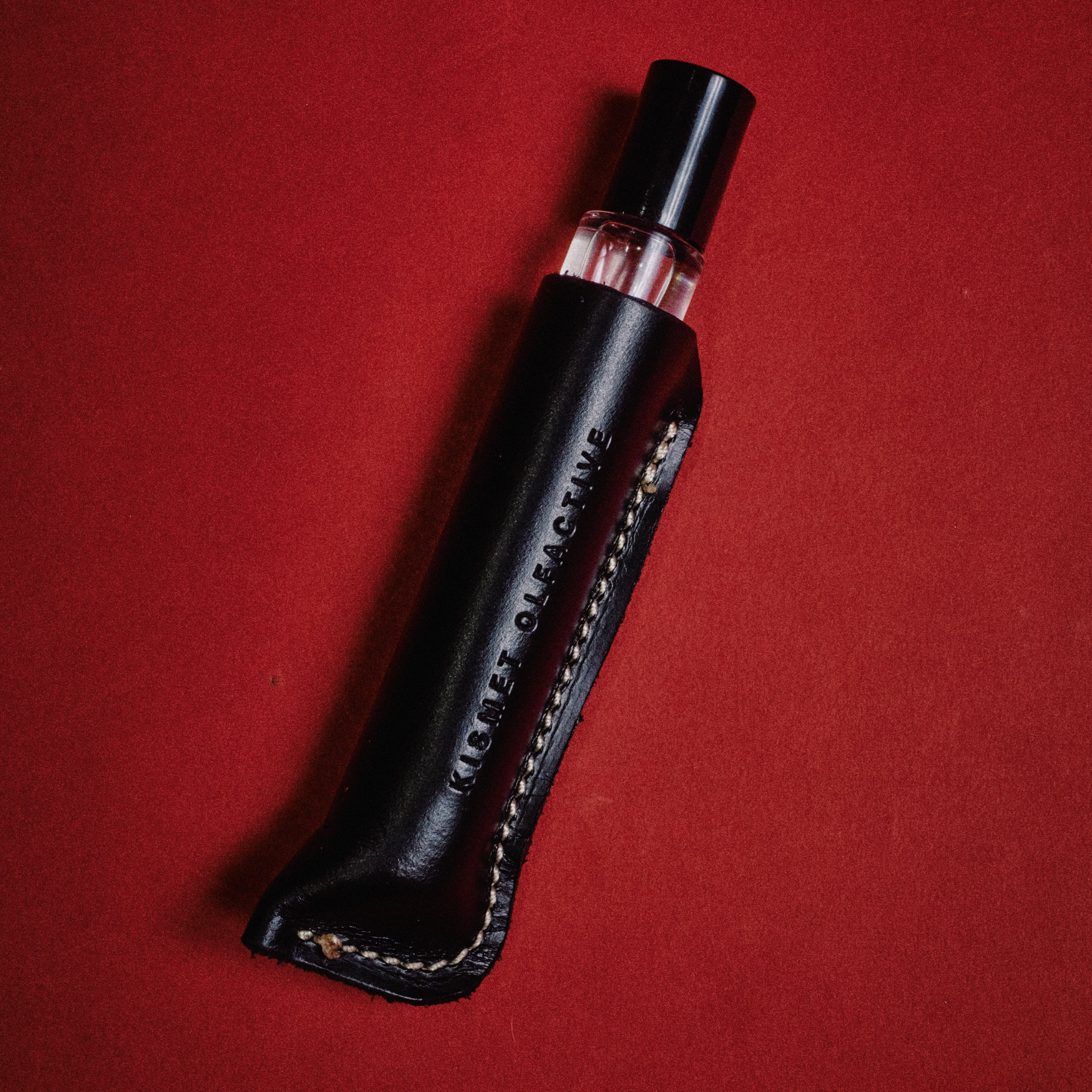 A black leather perfume bottle sleeve holding a clear glass perfume bottle with pink liquid, set diagonally on a textured red background. The sleeve features white contrast stitching and the brand name 'KISMET OLFACTIVE' debossed vertically.